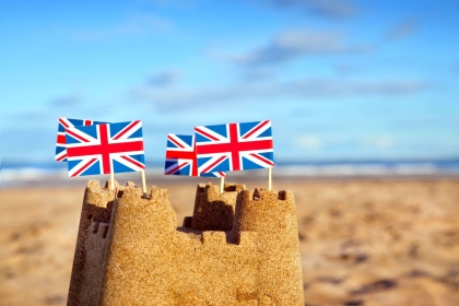 Sandcastle with Four Union Jack Flags Sticking out of Top. Pictured on Sunny Beach Underneath Blue Skies